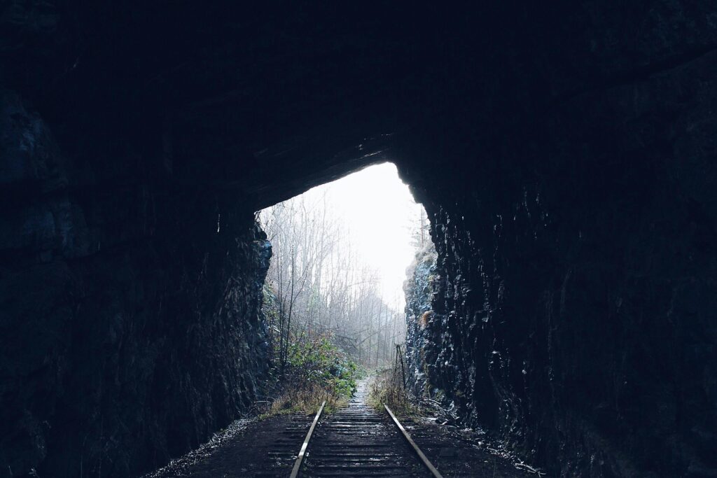 A dark railway tunnel with tracks leading to a bright natural landscape.